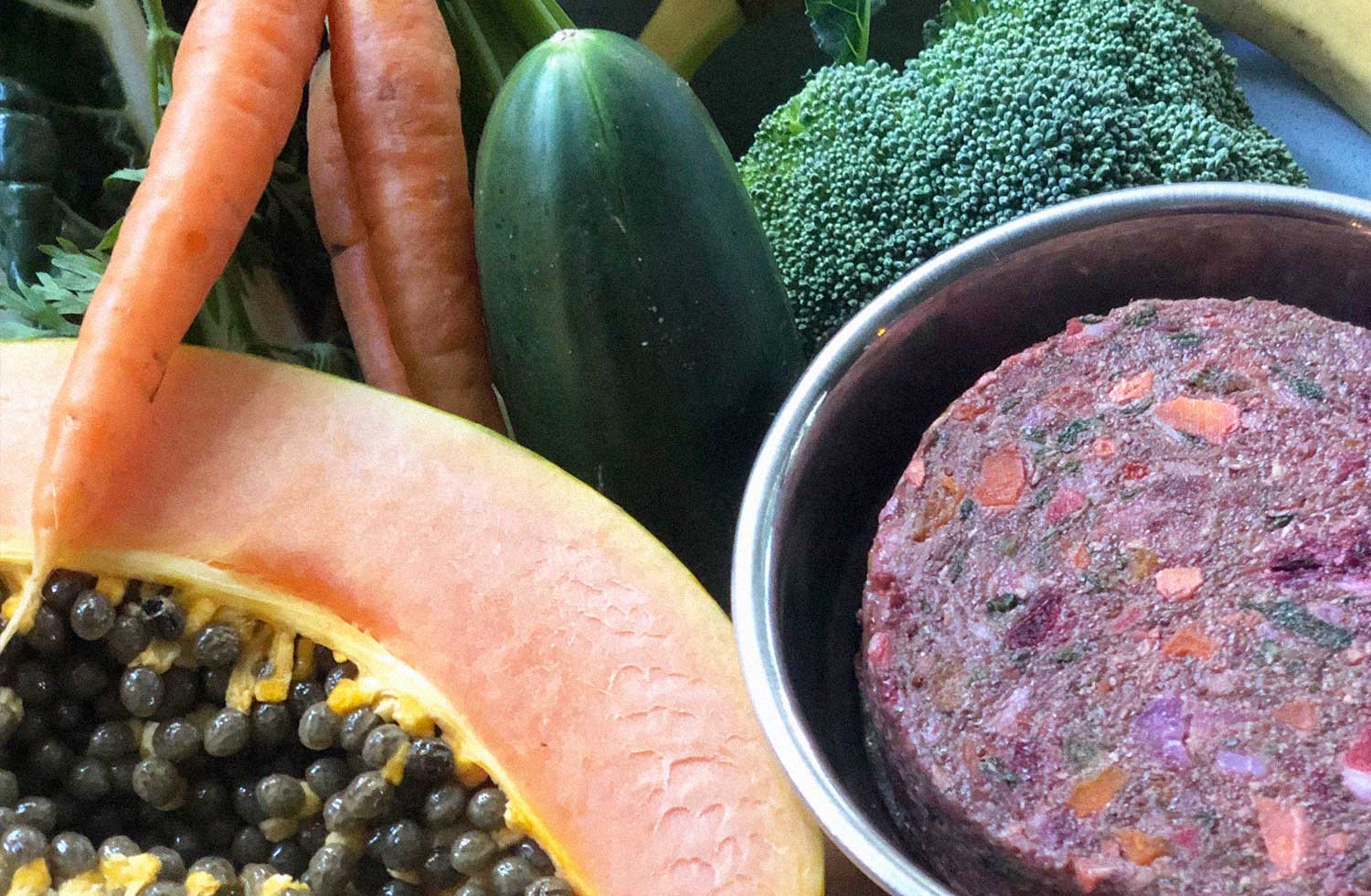 Butcher's Dog meal in a bowl with vegetables next to it including papaya, carrots, herbs and broccoli